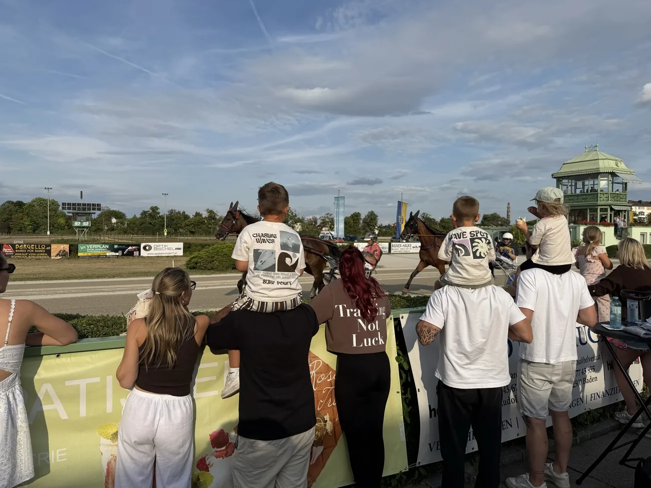 Children watching a harness horse race from the Baden racecourse finish straight.